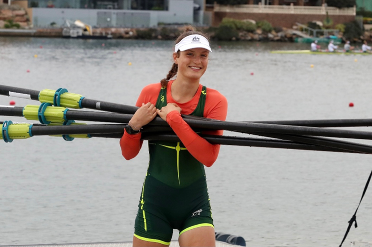 A photo of rower Gen Hart smiling at the camera and carrying rowing oars