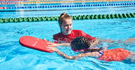 A photo of a swim instructor assisting a child in swimming using a kick board